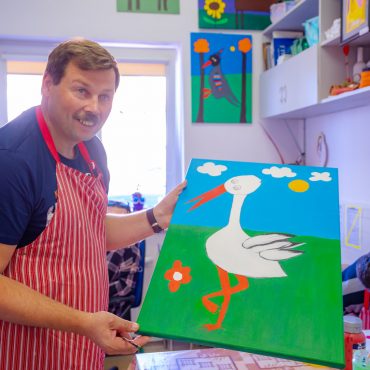 Mężczyzna w fartuchu w paski prezentuje obraz z namalowanym bocianem