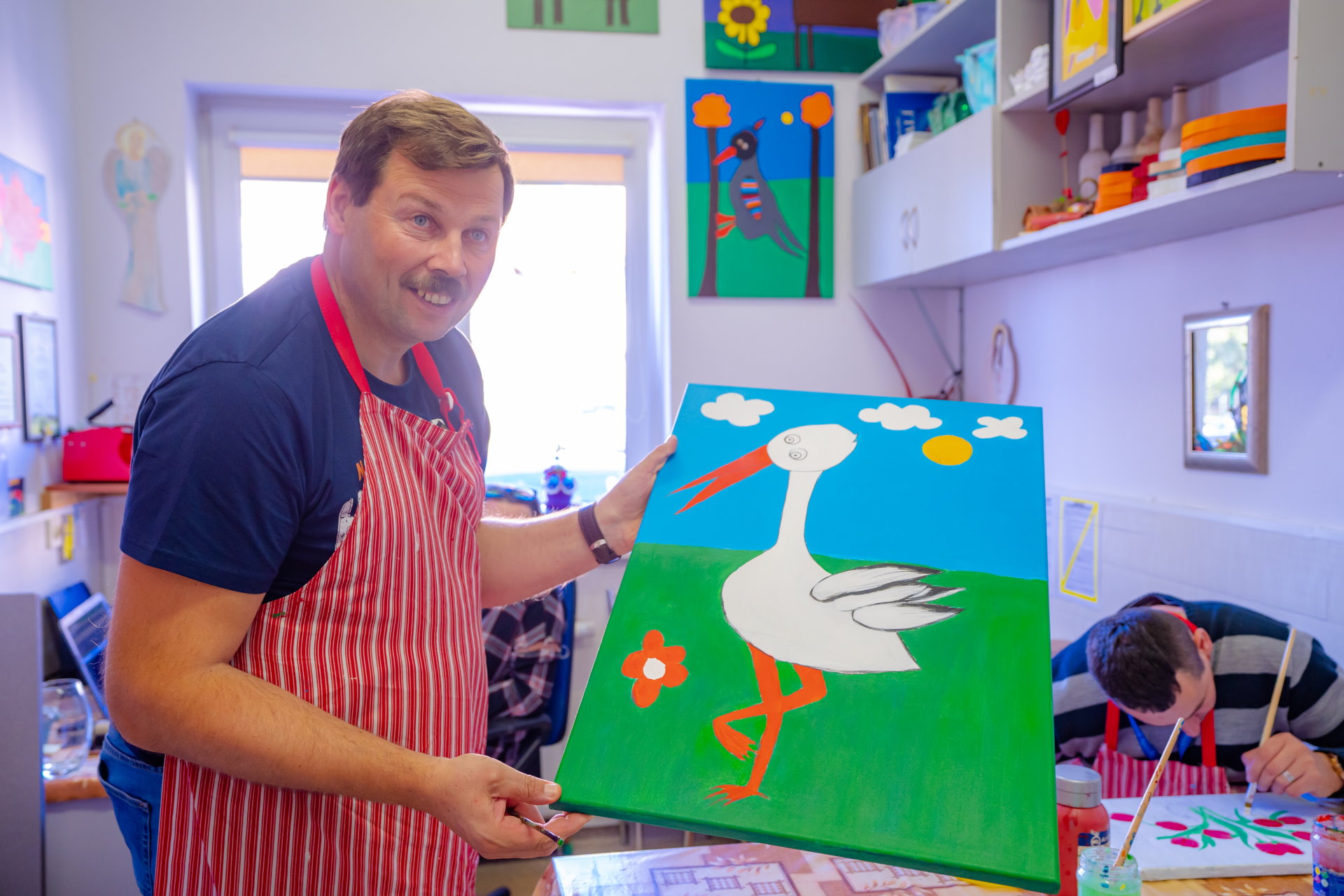 Mężczyzna w fartuchu w paski prezentuje obraz z namalowanym bocianem