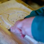 Osoba w zielonym kitlu trzyma ostre narzędzie i wykonuje drzeworyt