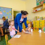 Trójka dzieci siedzi przy stole, nauczycielka w niebieskim swetrze kładzie na stole kartkę. Z tyłu siedzi kobieta przy laptopie