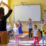 Kobieta podnosi ręce i łączy dłonie nad głową. Przed nia grupa dzieci, która naśladuje jej ruchy