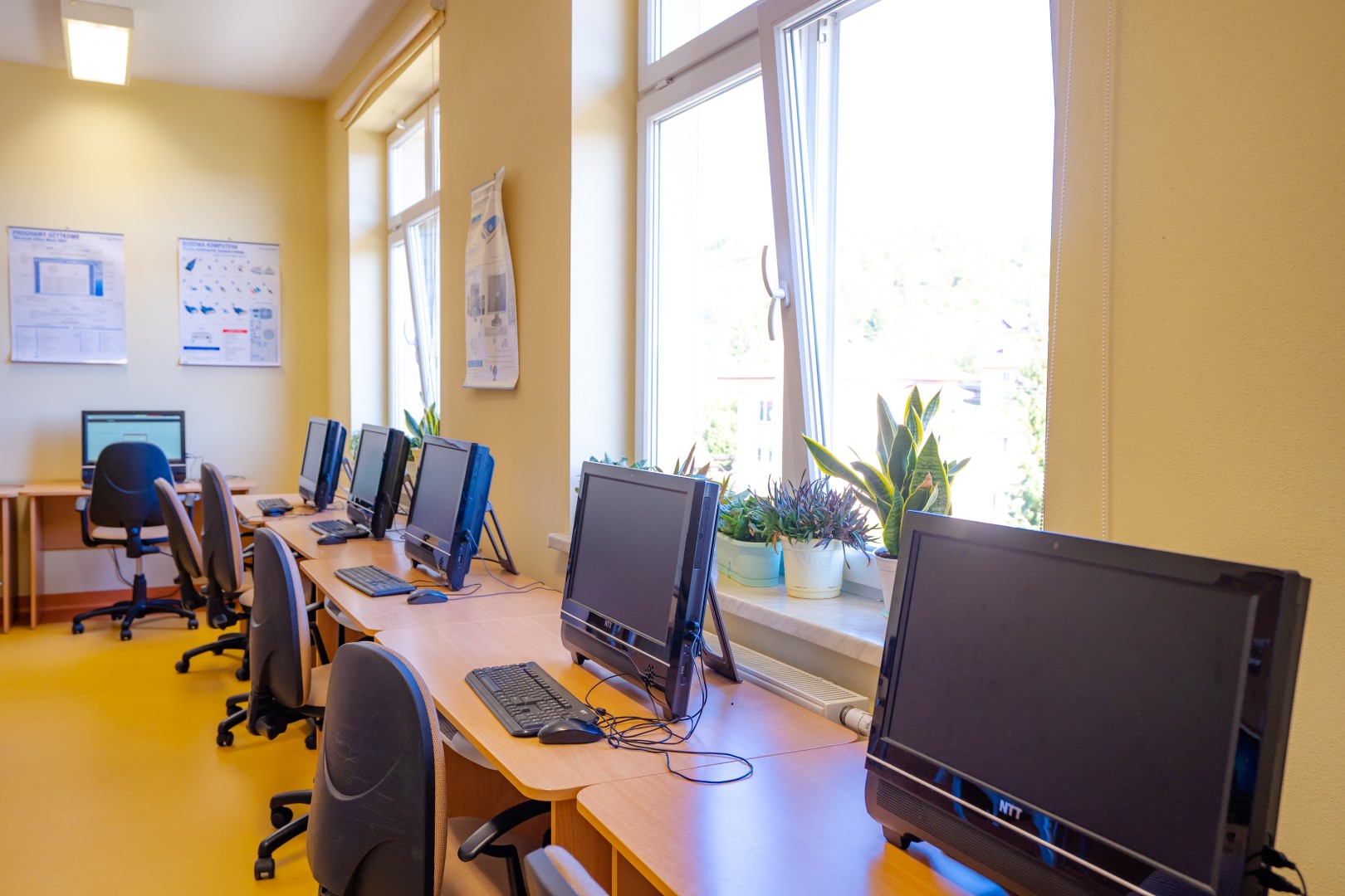 Wygaszone monitory koloru czarnego i klawiatury ustawione na biurkach. Przy biurkach fotele obrotowe