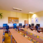 Klasa szkolena. Na środku stoły, a przy nich żółte krzesła. Pod ścianą biurka, a na nich monitory.