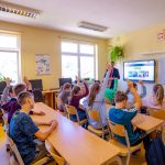 Lekca w klasie. nauczyciel w garniturze stoi pod ścianą. Uczniowie siedzą w ławkach i się zgłaszają