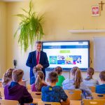 Lekcja w szkole. Nauczyciel w garniturze stoi przed ławkami. W ławkach siedza uczniowie.