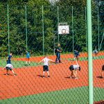 Cjłopcy w strojach sportowych ćwiczą na boisku. Boisko jest ogrodzone wysoką siatką.