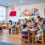 Dzieci siedzą przy małych stolikach. Nad stołem nachyla się kobieta