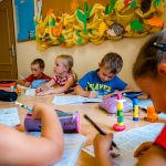 Dzieci w kolorowych bluzkach siedzą przy stole i piszą