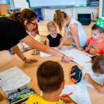 Nauczycielka w czarnej bluzce wskazuje coś w zeszycie. Przy stole dzieci piszą w zeszytach