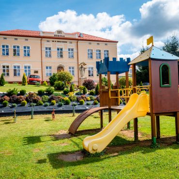 Plac zabaw wśród zieleni. W głębi różowy budynek ze spadzistym dachem