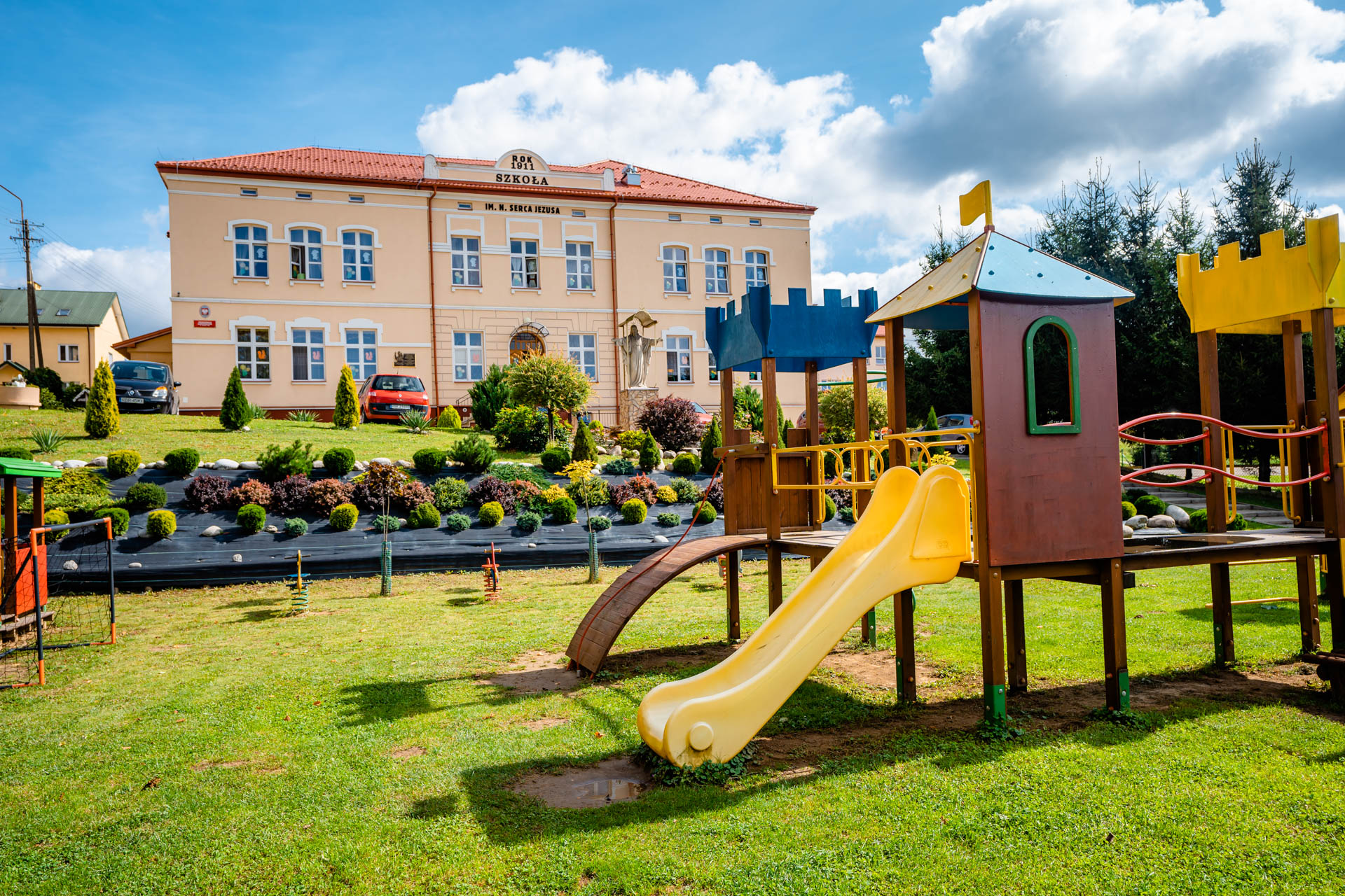 Plac zabaw wśród zieleni. W głębi różowy budynek ze spadzistym dachem