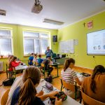 Uczniowie siedzą w ławkach w sali pomalowanej na kolor zielony. Na ścianie wiszą monitory interaktywne