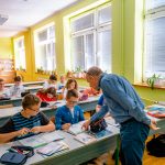 Nauczyciel pochyla się nad ławką, przy której siedzą uczniowie i coś objaśnia