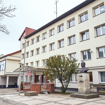 Czterokondygnacyjny budynek urzędu z jasną elewacją. Przed budynkiem popiersie i drzewa