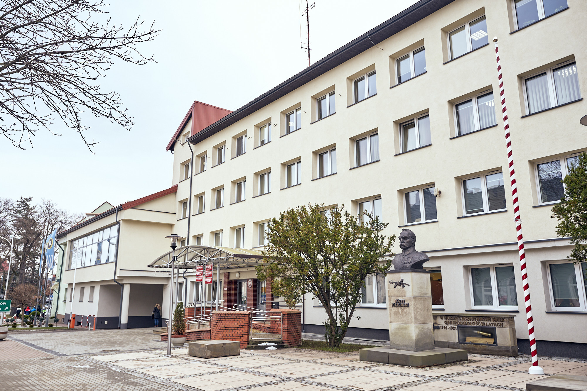 Czterokondygnacyjny budynek urzędu z jasną elewacją. Przed budynkiem popiersie i drzewa