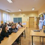 Kobieta z kręconymi bląd włosami pisze na cyfrowej tablicy. W ławkach siedzą uczniowie i patrzą się na tablicę