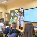 Kobieta z kręconymi bląd włosami stoi przy cyfrowej tablicy. W ławkach siedzą uczniowie i patrzą się na kobietę