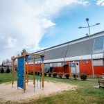 Bydynek hali sportowej z czerwoną elewacją. Przed halą plac zabaw