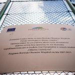 Srebrna metalowa tavlica z logotypami zawieszona na siatce ogrodzeniowej