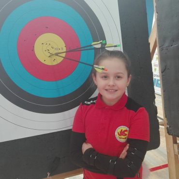Dziewczynka w stroju łuczniczki stoi przy tarczy z wbitymi strzałami