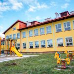 Dwukondygnacyjny budynek szkoły w kolorze żółtym pokryty czeerwonym spadzistym dachem. Przed budynkiem plac zabaw