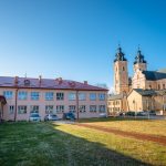 Dwukondygnacyjny budynek szkolny z czerwonym dachem. W głębi barokowy kościół z dwoma wieżami