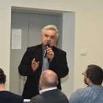 Siwy mężczyzna w czarnej marynarce mówi do mikrofonu