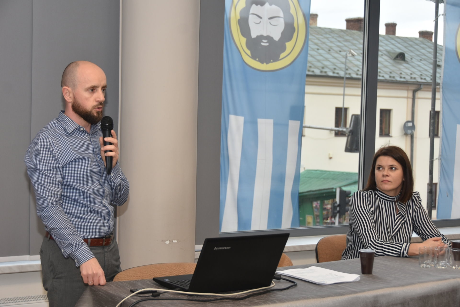 Łysiejący mężczyzna w koszule w kratę mówi do mikrofonu. Obok siedzi kobieta w bluzce w czarne paski