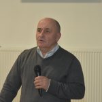 Łysiejący mężczyzna w szarym swetrze mówi do mikrofonu