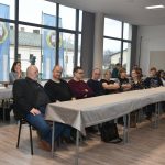 Grupa osób siedzi za długim stołem przykrytym obrusem. Z tyłu duże okna z widokiem na błękitno-białe flagi