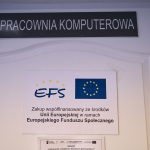 Białe drzwi, nad nimi czarna tablica z białym napisem. Na drzwiach tablica z flagą europejską i napisem EFS