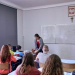 Klasa szkolna, uczniowie siedzą w ławkach. Nauczycielka pochyla się nad stołem. Na ścianie godło Polski i krzyż