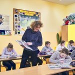 Nauczycielka z książką w ręce patrzy na dzieci siedzące w ławkach i odrabiające zadanie