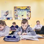 Dzieci siedzą w ławkach i pisza w zeszytach. Z tyłu na ścianie wisi mapa Polski