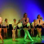 Dziewczyny w białych bluzkach i czarnych krótkich spodeńkach tańczą na scenie