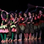 Grupa dzieci w kolorowych strojach i dużych czerwonych kapeluszach występuje na scenie