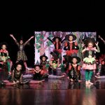 Grupa dzieci w kolorowych strojach i dużych czerwonych kapeluszach występuje na scenie