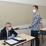 Mężczyzna w garniturze siedząc przy stoliku zadaje pytanie. Z kolei mikrofon trzyma mu młody wysoki mężczyzna.