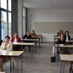 Grupa osób w tym mężczyzn i kobiet słuchają i ogladają wideokonferencji.