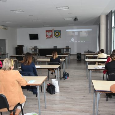 Grupa osób w tym mężczyzn i kobiet słuchają i ogladają wideokonferencji.