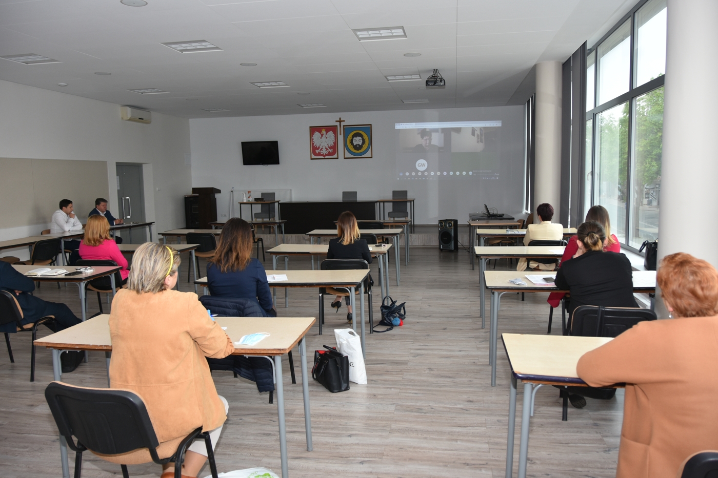 Grupa osób w tym mężczyzn i kobiet słuchają i ogladają wideokonferencji.