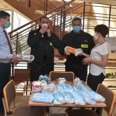 Mężczyna przekazuje maseczki dla dwóch strażaków w asyście kobiety w holu budynku