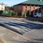 Przejście dla pieszych z progiem zwalniającym. Po drugiej stronie ulicy budynek koloru brązowego ze spadzistym zielonym dachem.