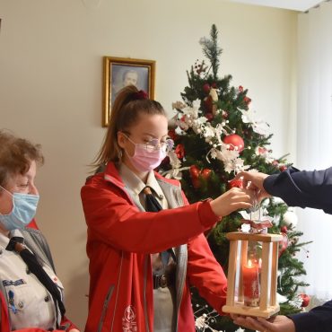 Młody mężczyzna odbiera od dwóch kobiet lampion z zapaloną świecą. W pokoju stoi też choinka.
