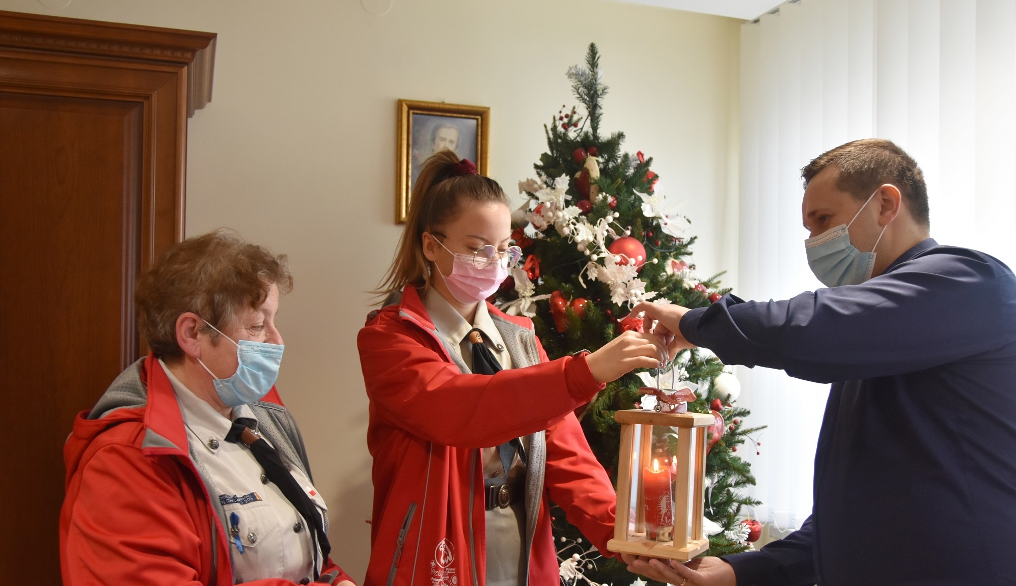 Młody mężczyzna odbiera od dwóch kobiet lampion z zapaloną świecą. W pokoju stoi też choinka.