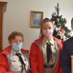 Dwie kobiety w garniturach i mężczyzna w koszuli przed choinką pozują do zdjęcia. Ponadto młody mężczyzna trzyma w ręku lampion z zapaloną świeczką.