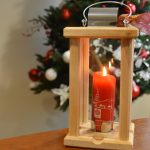 Szklany lampion z drewnianym wykończeniem, zapaloną świecą znajduje się na stole w pokoju z choinką.