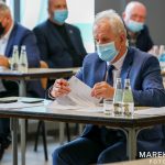 Starszy mężczyzna w maseczce siedzi przy stoliku czytając dokumenty. W tle widać trzech mężczyzn w średnim wieku w garniturach.