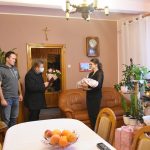 Starszy mężczyzna w ciemnym płaszczu składa gratulacje, przekazując kwiaty młodej dziewczynie, która trzyma na rękach malutkie dziecko w białym rożku. W pokoju znajdują się też młody mężczyzna i kobieta w średnim wieku.