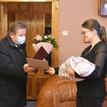 Starszy mężczyzna w ciemnym płaszczu przekazuje kwiaty młodej kobiecie trzymającej na rękach dziecko w rożku.